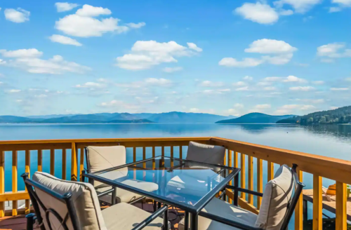 A scenic view of a calm lake from a wooden deck with a glass table and chairs, under a blue sky with fluffy clouds.